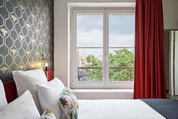 A hotel room with a bed, a window with a red curtain, a view of the city, and a patterned wall.