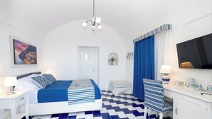 A hotel room with a checkered floor, a bed with blue and white bedding, a desk with a lamp, a chair with a blue and white pattern, and a TV on the wall. - B&B Casa Nilde Positano (Positano)