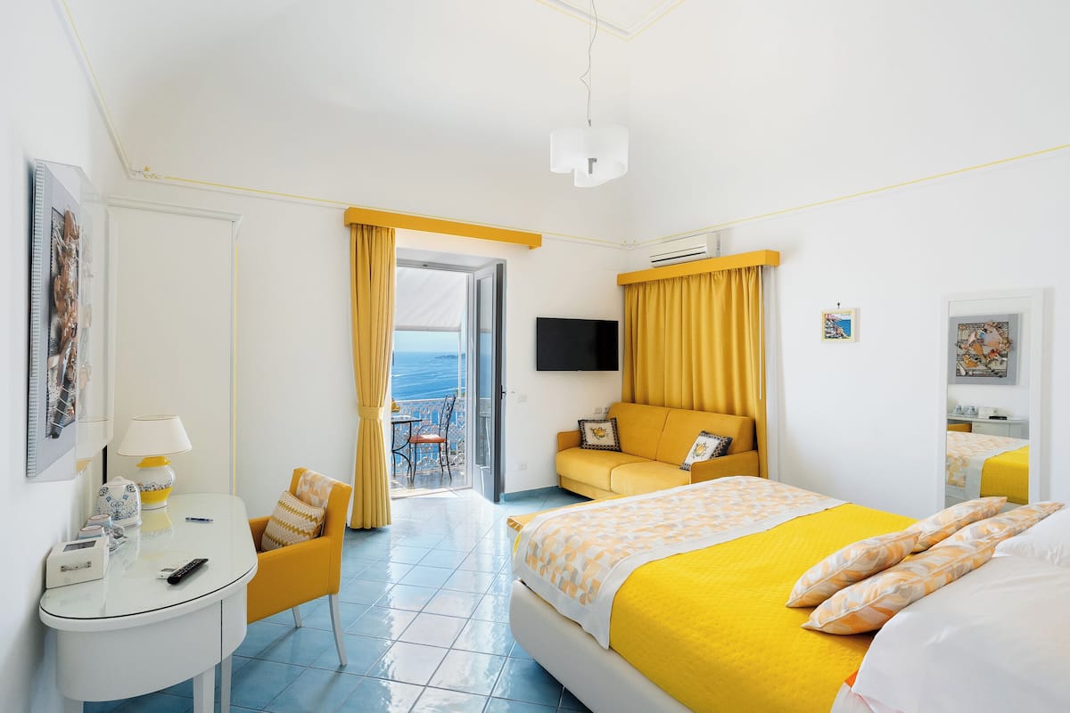 A hotel room with a bed, a desk, a chair, a TV, and a balcony with yellow curtains.