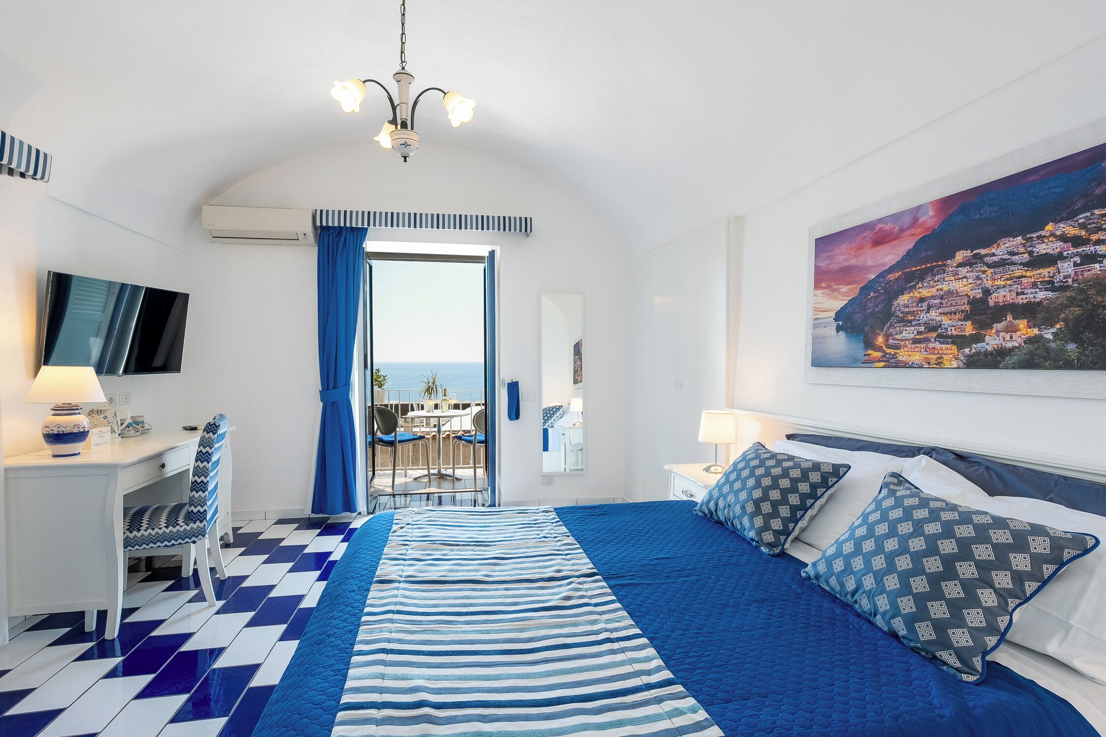 A bedroom with a checkered floor, a bed with blue and white bedding, a desk with a chair, a TV, and a painting of a coastal town.
