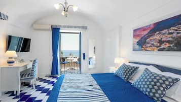 A bedroom with a checkered floor, a bed with blue and white bedding, a desk with a chair, a TV, and a painting of a coastal town.