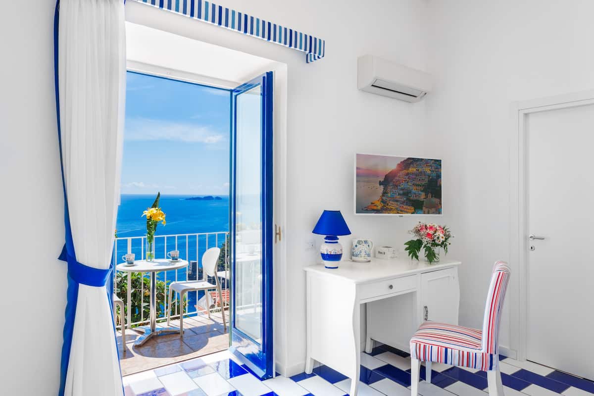 A room with a balcony, a desk, a chair, and a painting on the wall.