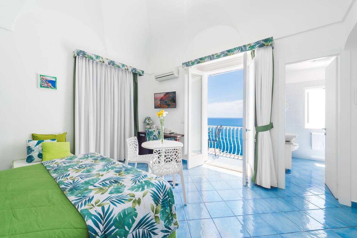 A hotel room with a bed, a small table with chairs, a view of the sea, and a bathroom.