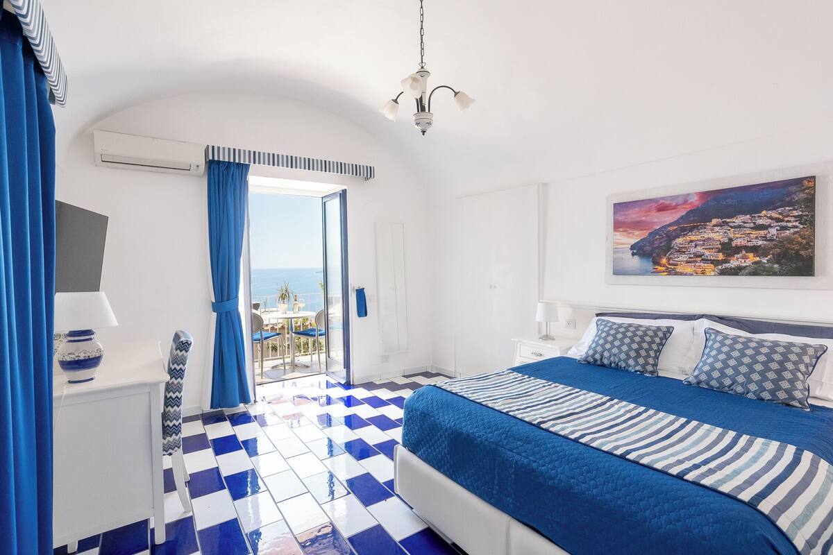 A bedroom with a checkered floor, a bed with blue and white bedding, a painting of a coastal town, and an air conditioning unit.