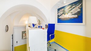 A reception area with a white desk, a blue chair, and a painting of a coastal scene. - B&B Casa Nilde Positano (Positano)