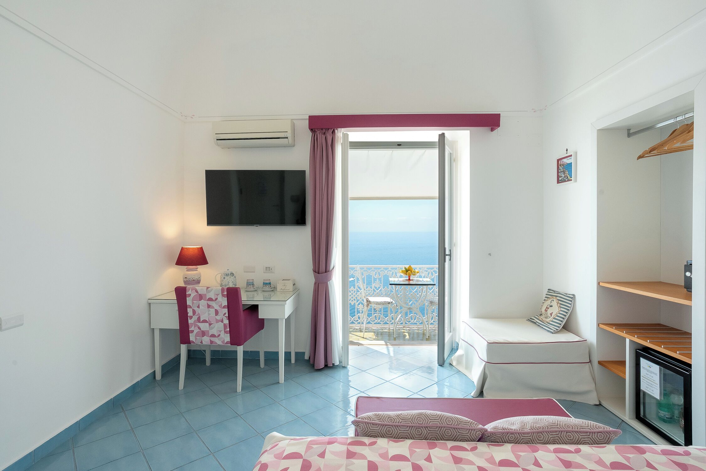 A hotel room with a bed, a desk, a chair, a TV, a balcony with a view of the sea, and a refrigerator.