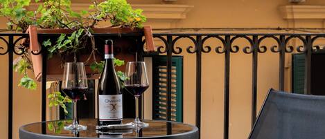 A round table with two glasses of red wine and a bottle, set against a balcony railing with a potted plant.
