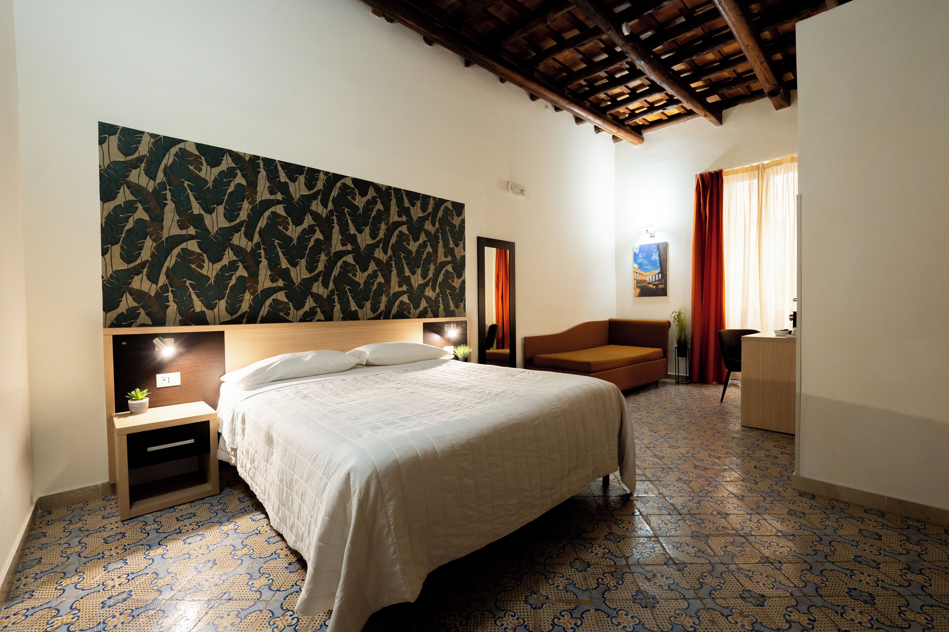 Une chambre à coucher comprenant un lit, une table de chevet, un bureau, une chaise, un sol à motifs et un plafond avec des poutres apparentes.