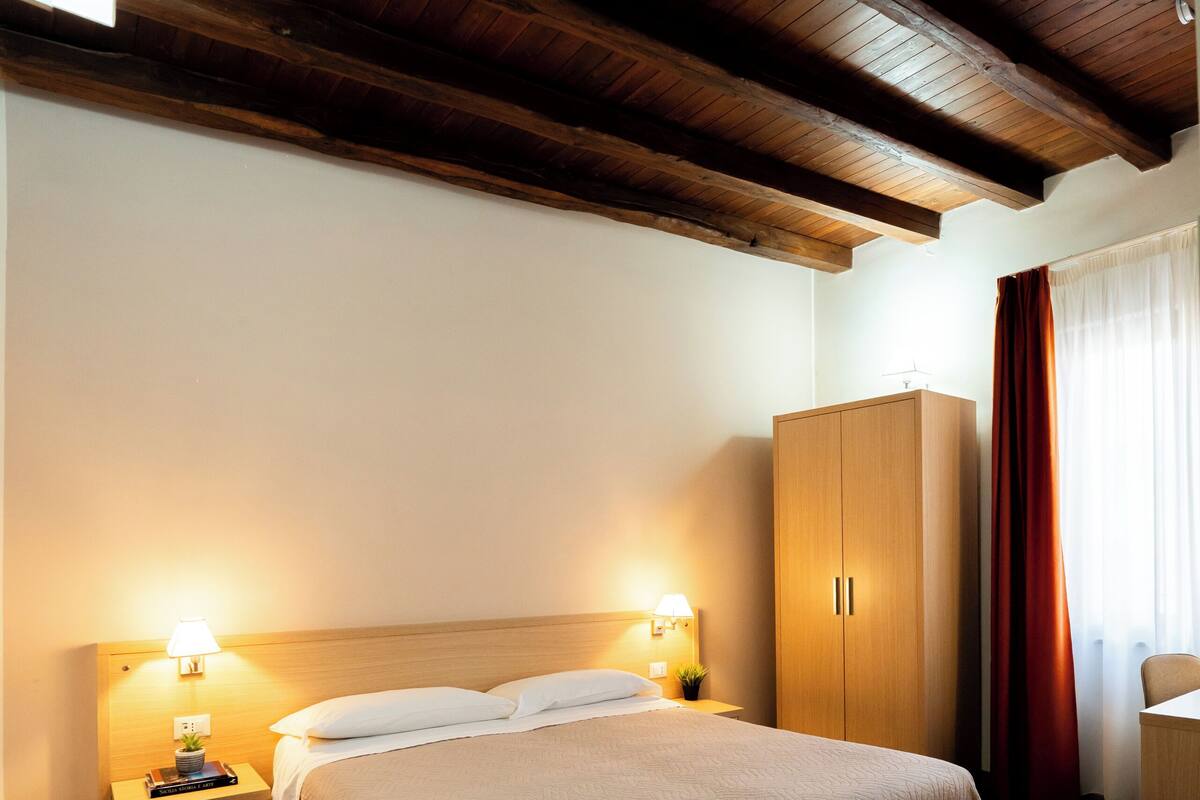 A hotel room with a wooden ceiling, a bed with a beige bedspread, a nightstand with a lamp, a wardrobe, and patterned floor tiles.