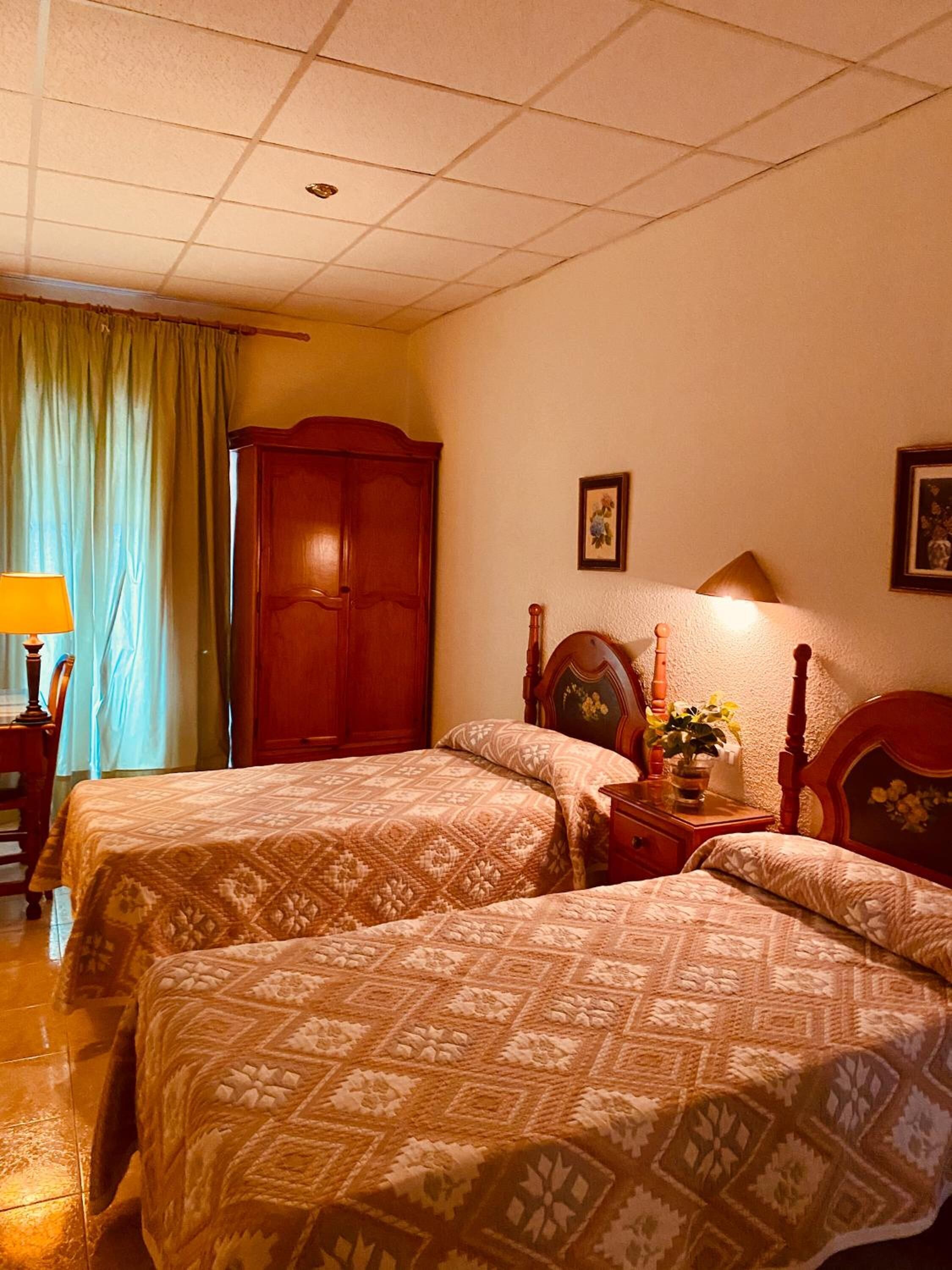 Quarto de hotel com duas camas, um guarda-roupa de madeira, uma mesa de cabeceira com abajur e uma janela com cortinas.