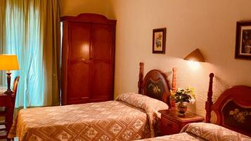 Quarto de hotel com duas camas, um guarda-roupa de madeira, uma mesa de cabeceira com abajur e uma janela com cortinas.