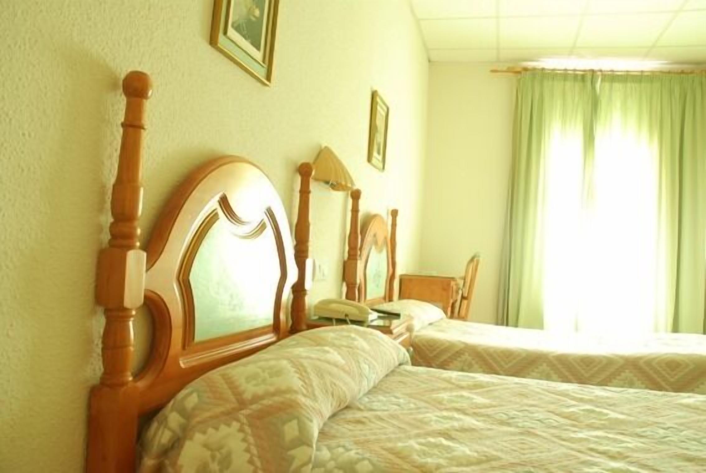 Een hotelkamer met twee houten bedden, een nachtkastje, een lamp en een raam met gordijnen.