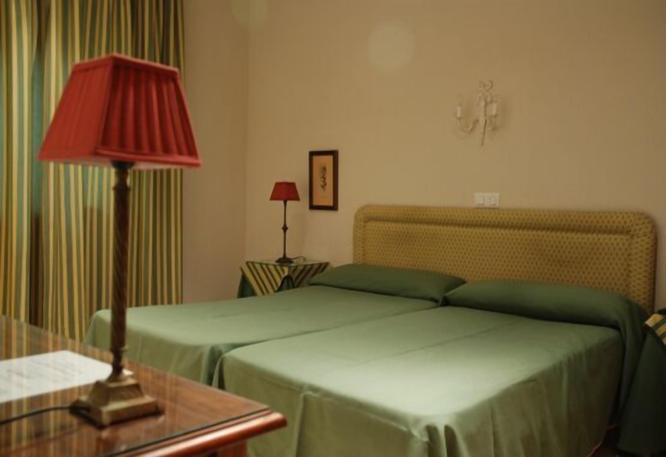 A hotel room with two beds, a wooden desk, a red lamp, a framed picture, and a patterned wall.