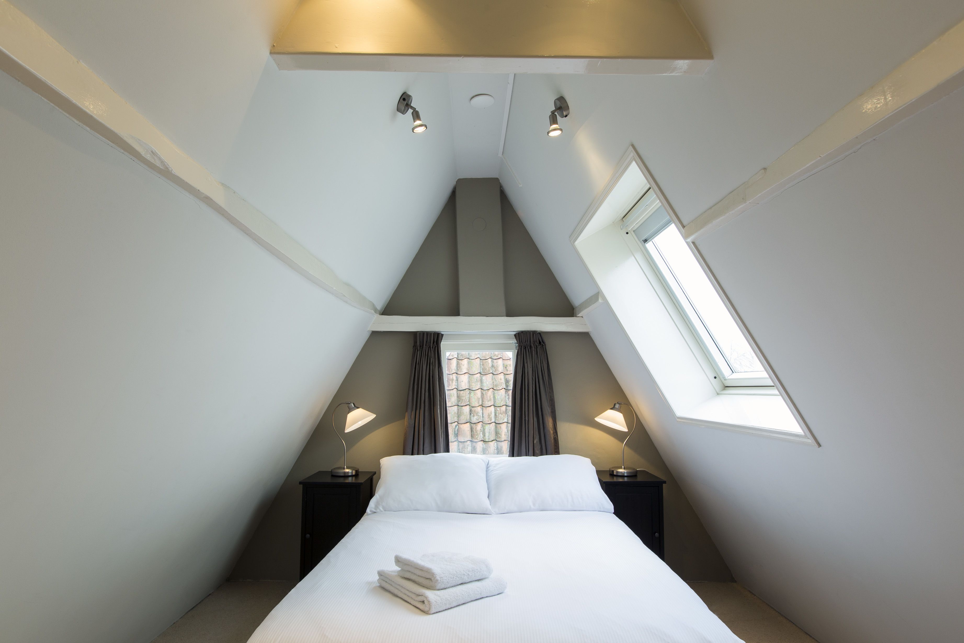 A bedroom with a sloped ceiling, a skylight, a bed with white linens, two bedside tables with lamps, and a window with curtains.