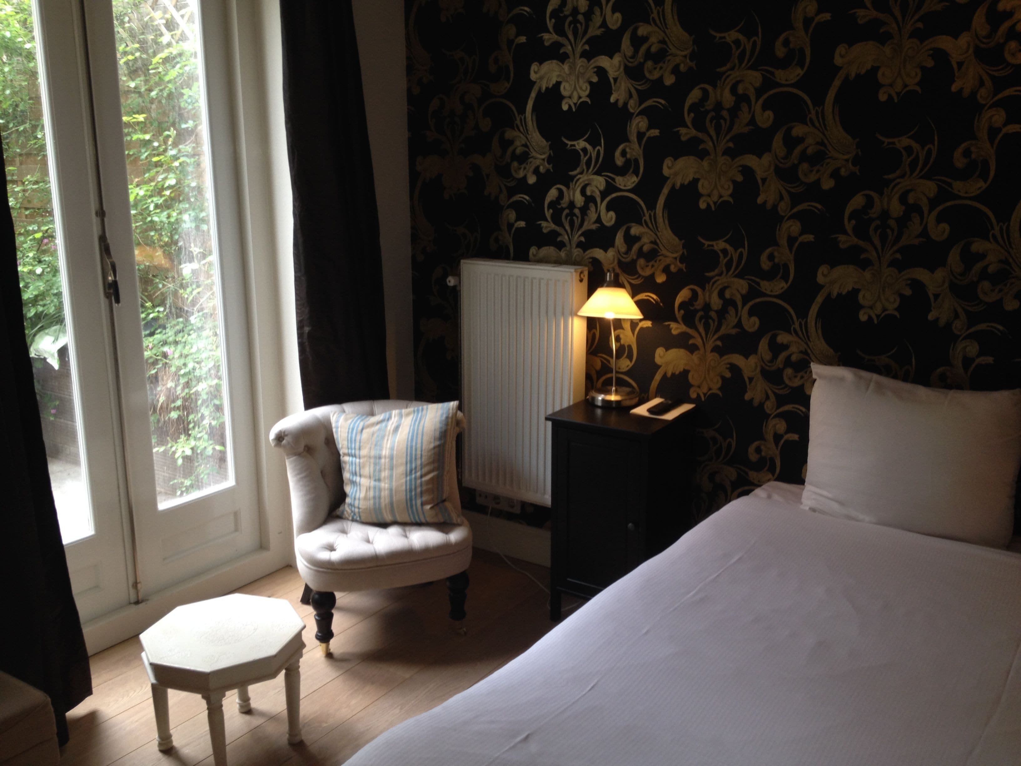 Une chambre à coucher comprenant un lit, une chaise, une table de chevet avec une lampe, un radiateur et une fenêtre avec des rideaux.