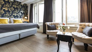 Un dormitorio con cama, dos sillones, una mesita con un jarrón y una copa de vino, y ventanas grandes con cortinas.