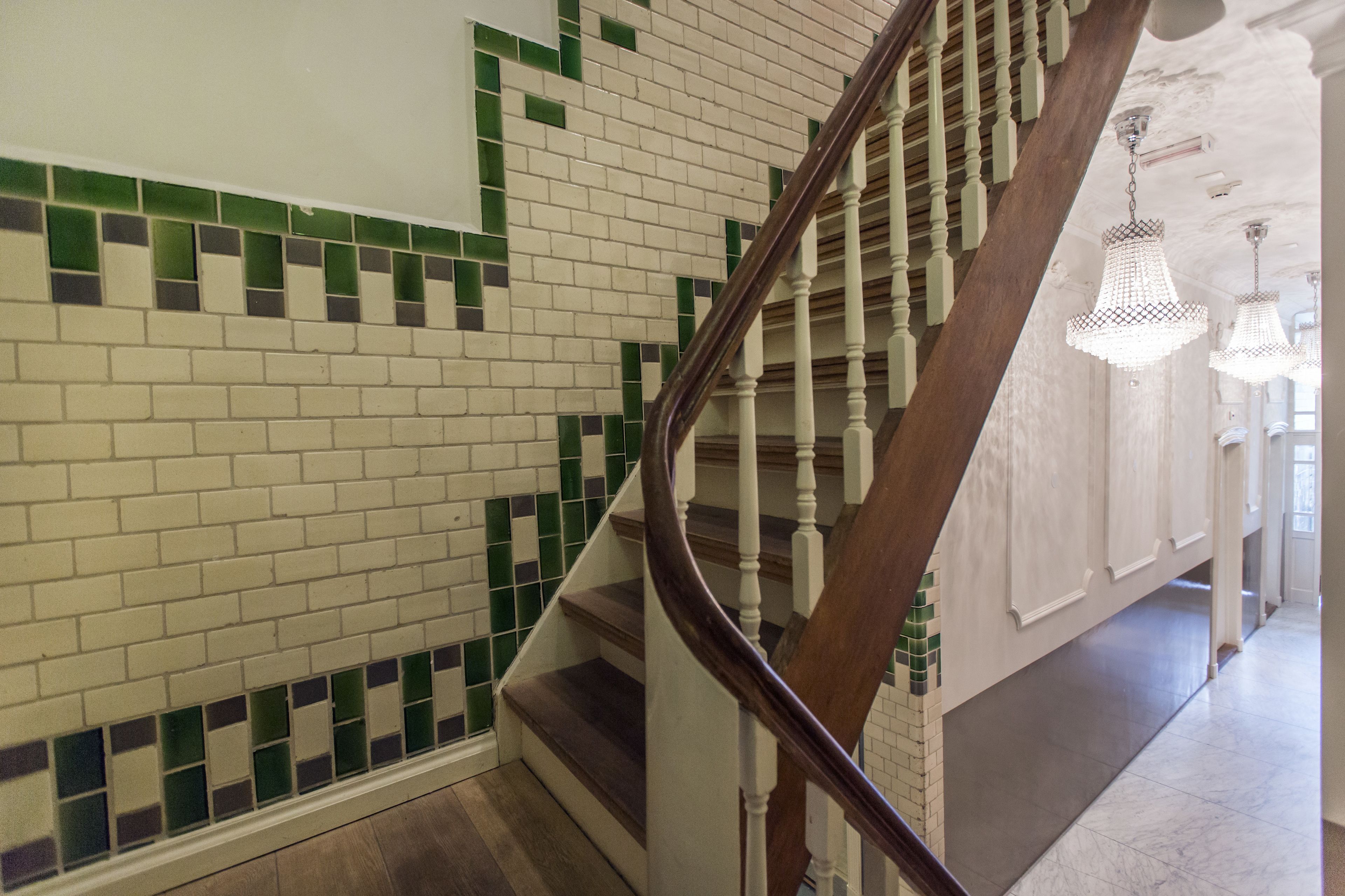 Una escalera con barandilla de madera y una pared blanca con baldosas verdes y beige.