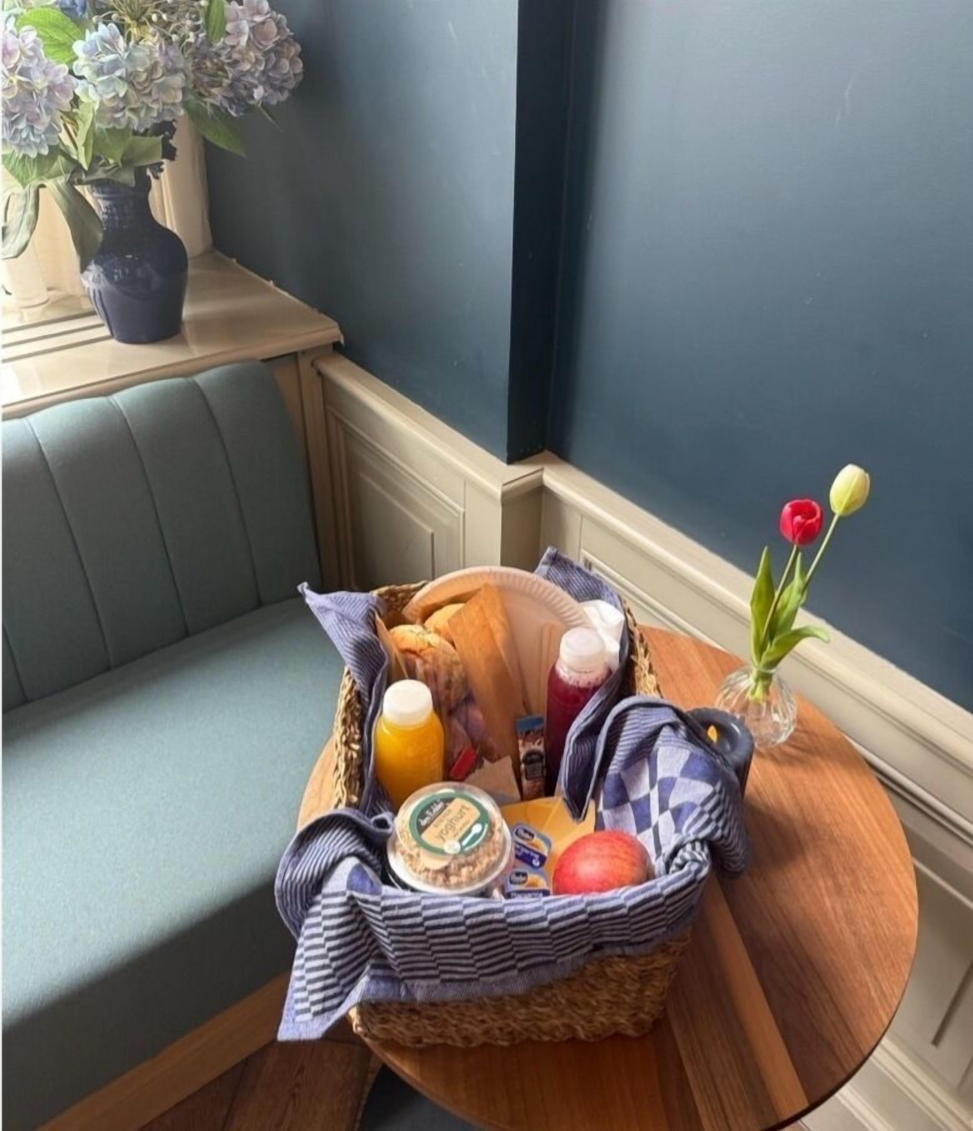 Una cesta de desayuno con pan, fruta y salsas sobre una mesa de madera.