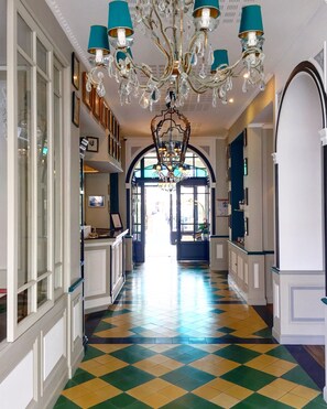 A hallway with a checkered floor, a chandelier, and framed pictures on the walls. - Hôtel Villa-Lamartine (Arcachon)