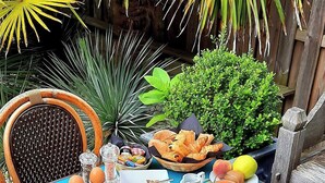 Mesa de café da manhã montada ao ar livre, com uma cadeira de vime, uma cesta de pão, um prato de presunto, ovos e um jarro de suco de laranja.
