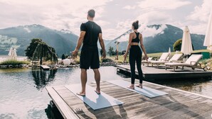 A wooden deck with two people standing on yoga mats, overlooking a body of water and mountains.