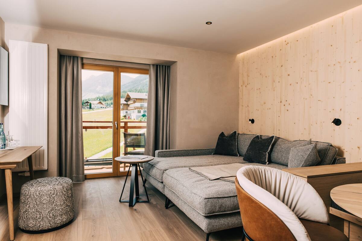 Una habitación de hotel moderna con un sofá, una pequeña mesa redonda, piso de madera y vistas a un paisaje montañoso por la ventana.