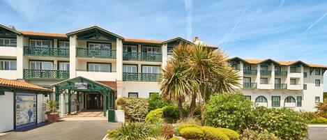 A hotel with a central entrance, multiple balconies, and a landscaped garden with various plants.