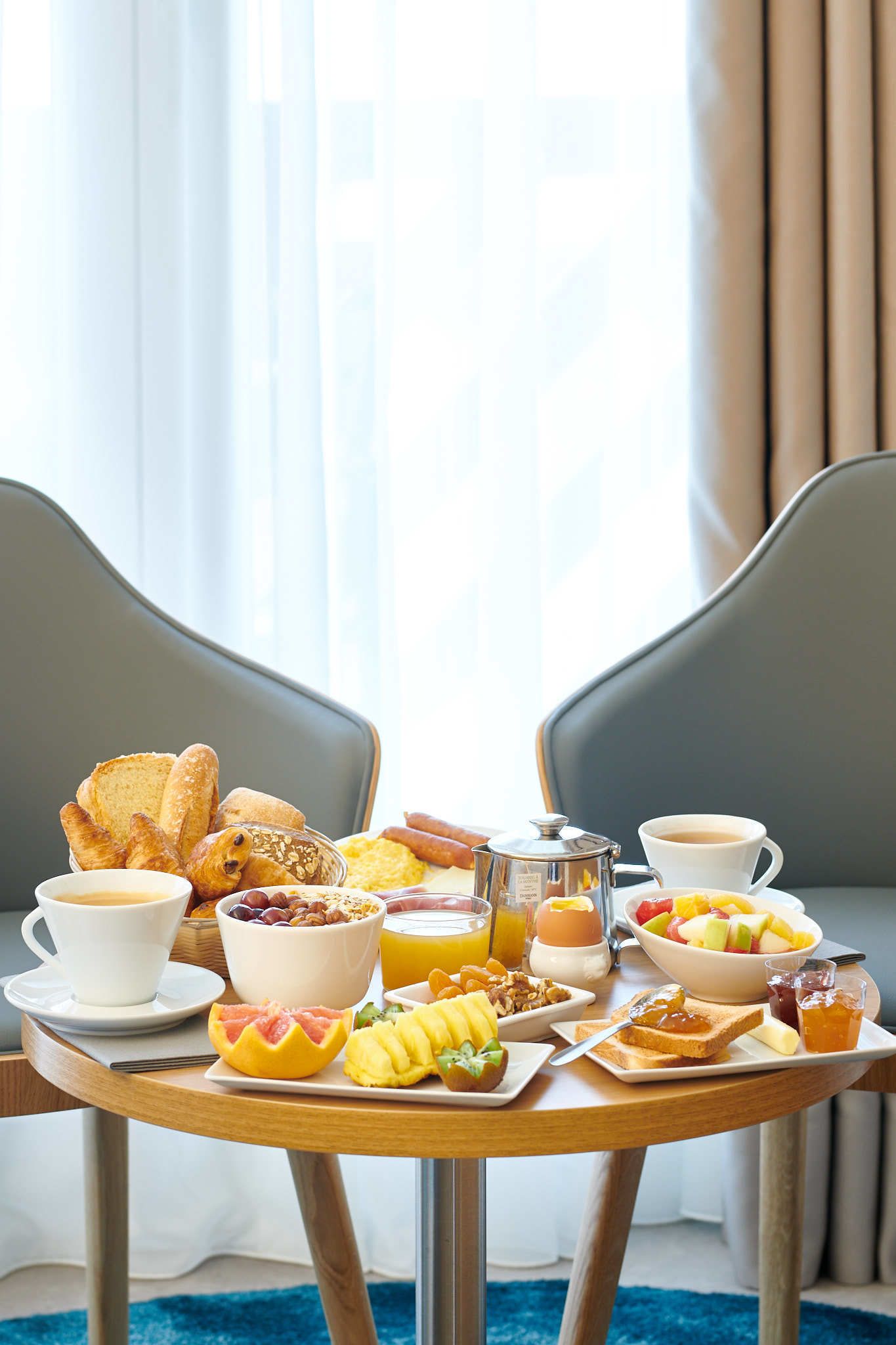 A breakfast table with various food items and beverages, set in a room with a view of curtains and a window.