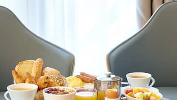 A breakfast table with various food items and beverages, set in a room with a view of curtains and a window.