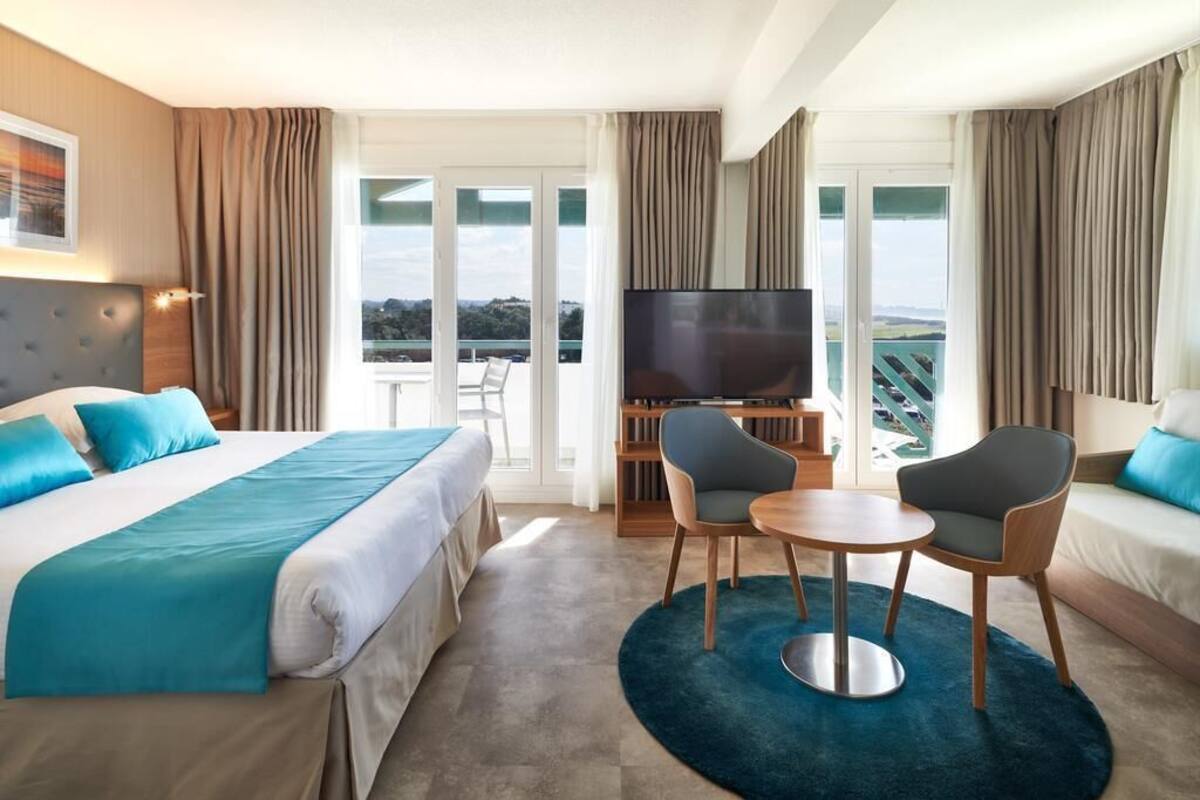 A hotel room with a bed, a TV, a balcony, and a seating area with a table and chairs.