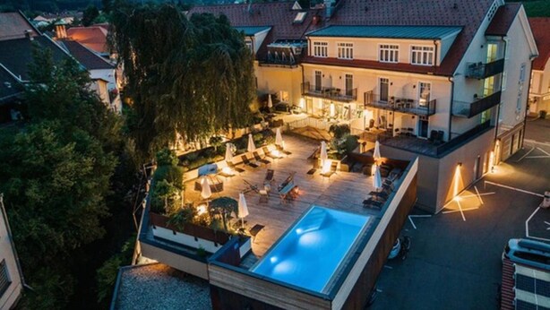 Un área de piscina en la azotea con sillones de descanso y sombrillas, rodeada de edificios y árboles.