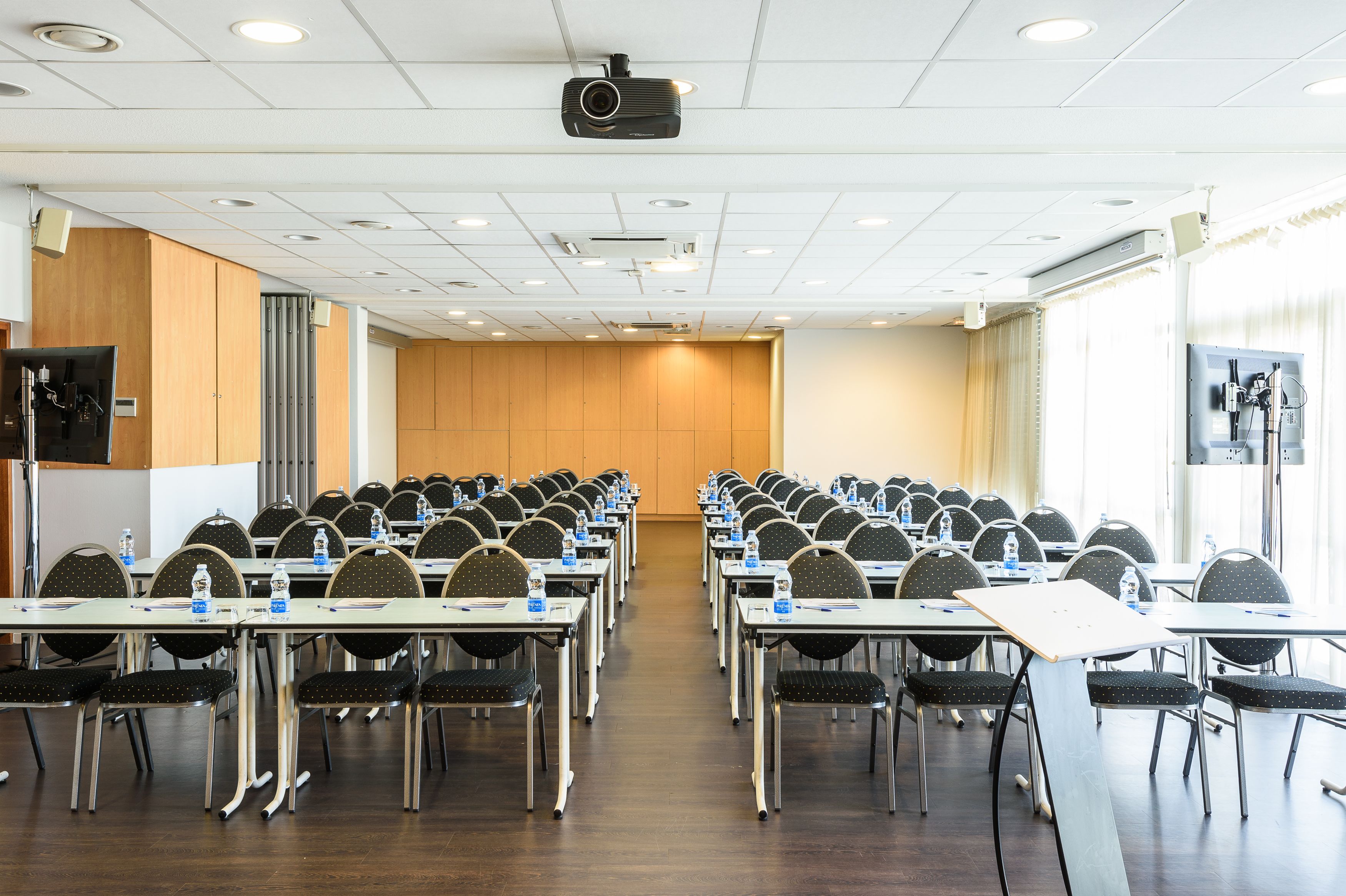 A conference room with rows of tables and chairs, equipped with projectors and water bottles.