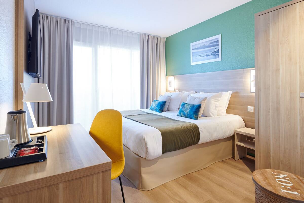 A hotel room with a bed, bedside table, lamp, chair, and a view of the outdoors through the window.