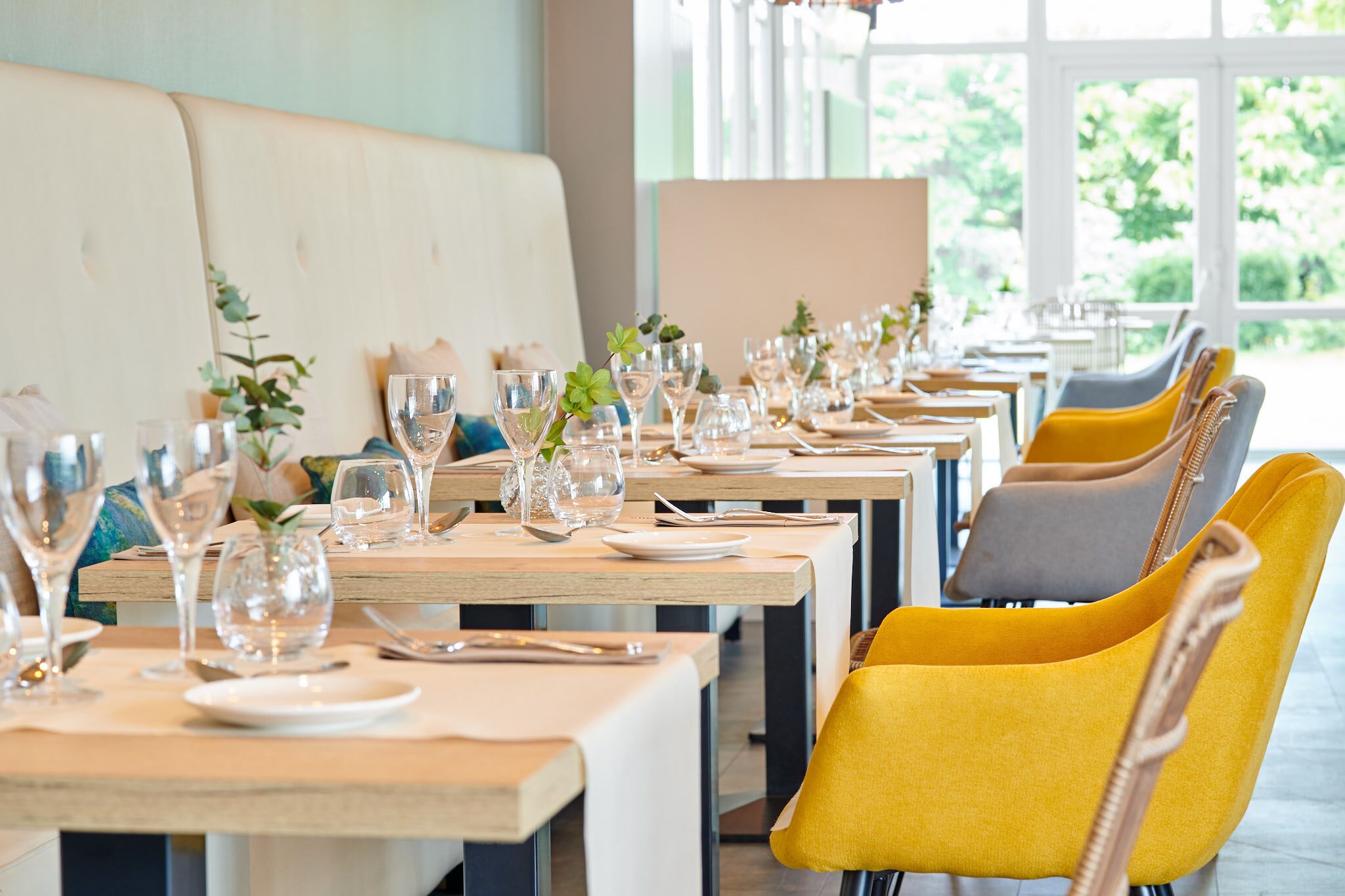 Un restaurante con mesas de madera largas, sillas amarillas y grises, y vasos de vidrio transparente.