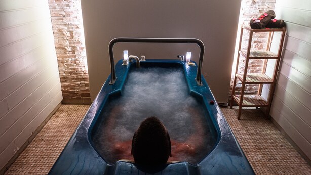 A person in a hot tub with a metal frame and faucets, surrounded by tiled walls and a wooden ladder with towels. - Hôtel Les Terrasses d'Atlanthal (Anglet)