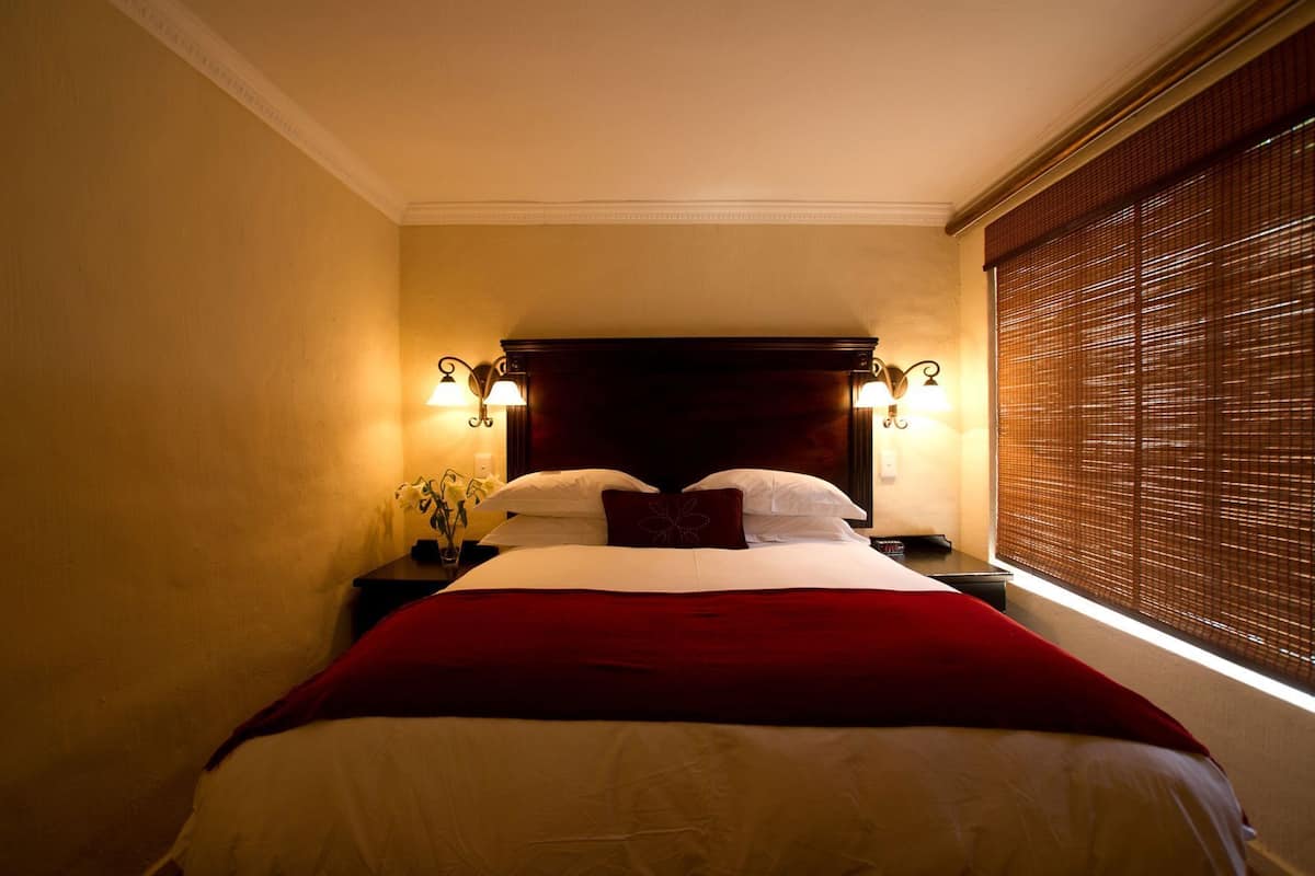 Une chambre d’hôtel avec un grand lit, deux tables de chevet, une petite table, une lampe et une fenêtre avec des stores.