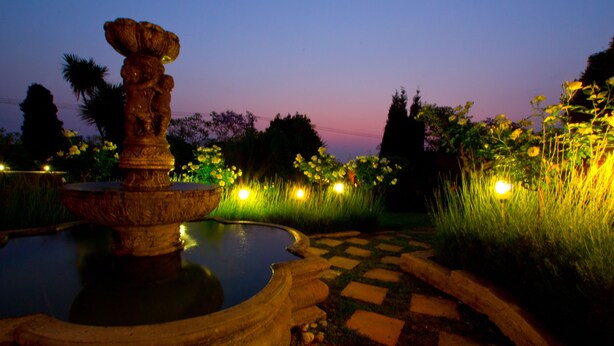 A fountain with a statue, surrounded by a garden with illuminated flowers and a stone pathway. - Little Tuscany Boutique Hotel (Sandton)