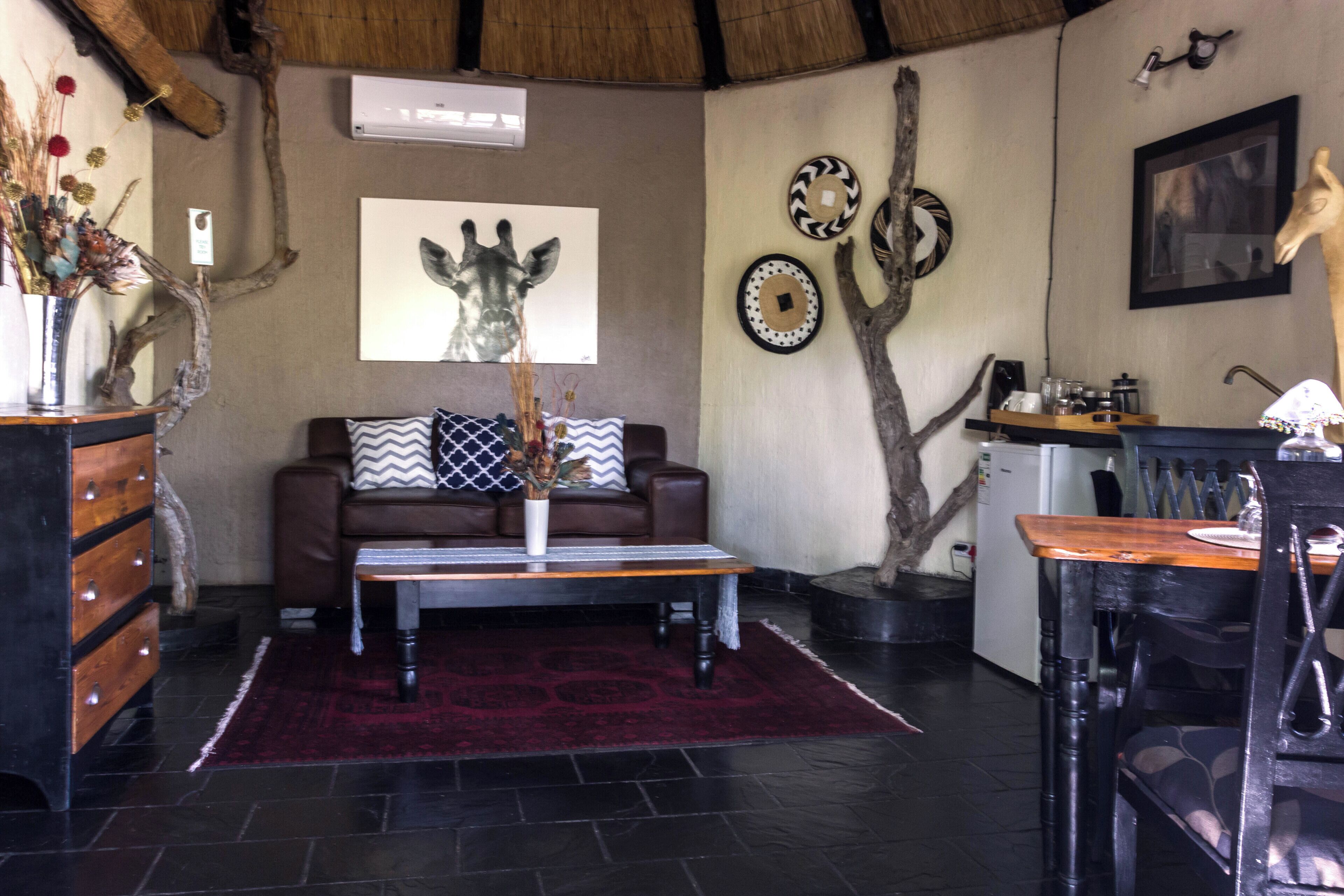 Un salon chaleureux avec un canapé en cuir marron, une table basse en bois et un tapis rouge. On y trouve un climatiseur fixé au mur, un tableau encadré représentant une girafe et des assiettes décoratives.