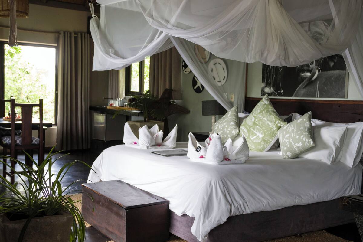 A neatly made bed with white linens and decorative pillows, a wooden bench, and a potted plant.