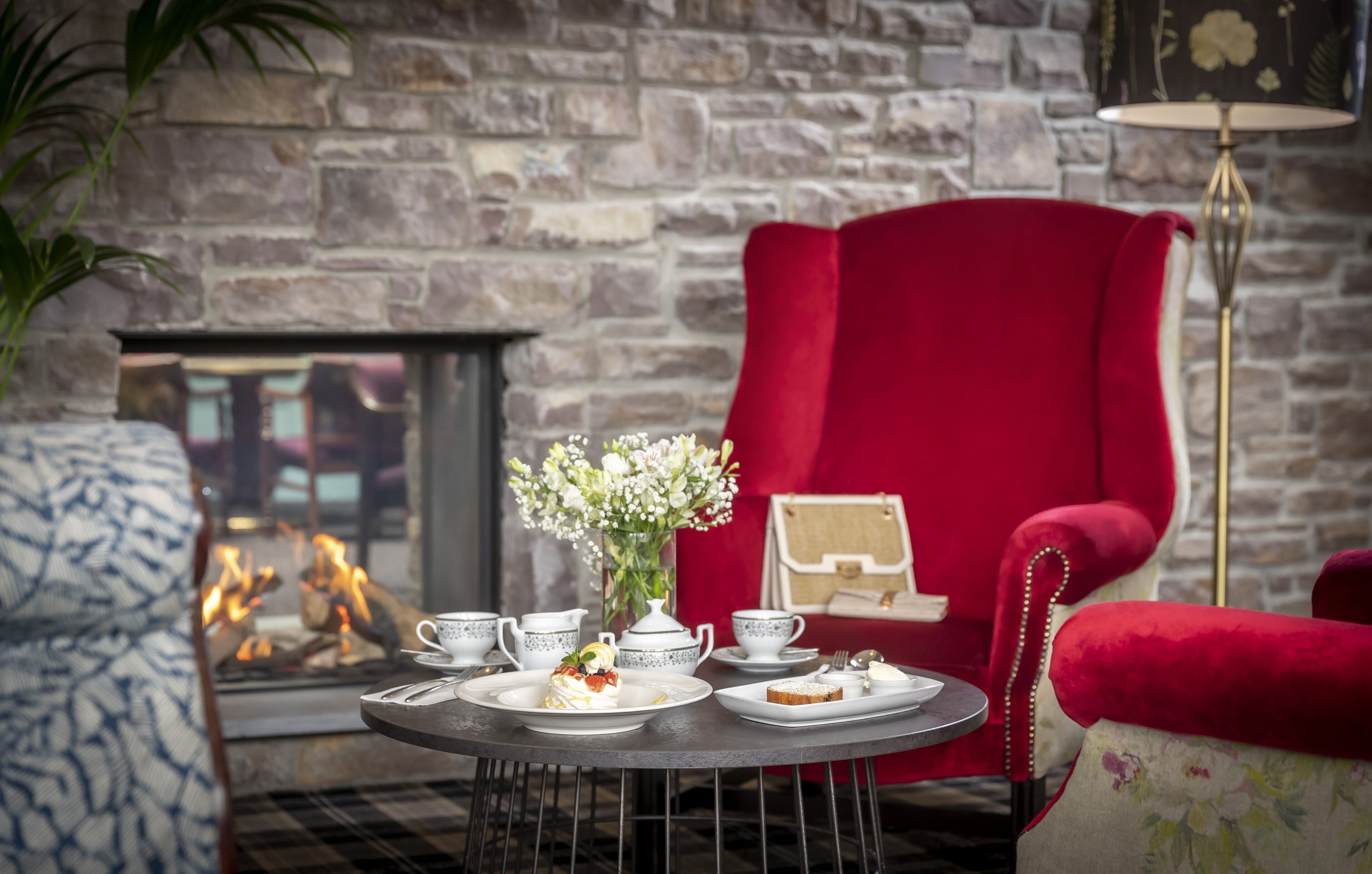 A cozy room with a fireplace, a red armchair, a round table with tea service, and a patterned sofa.