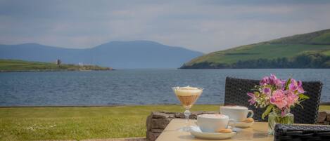 A table with a glass of a layered beverage, two white cups, and a vase with pink flowers, set against a scenic view of a lake and hills.