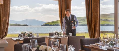 A restaurant with tables set for dining, a waiter attending to guests, and a scenic view of a lake and hills.