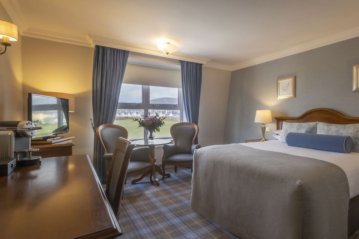 A hotel room with a bed, two chairs, a desk with a television, and a window with curtains.