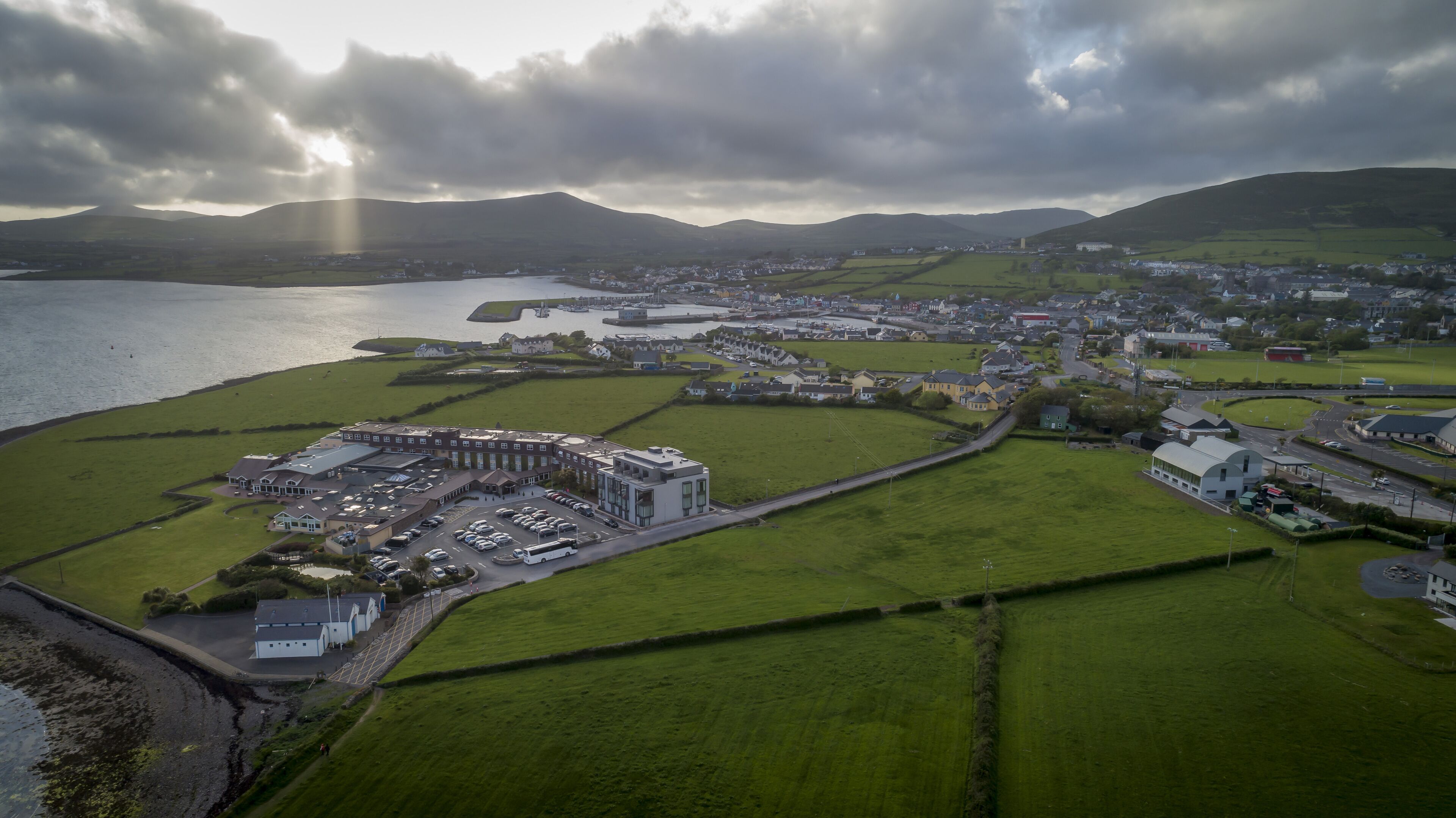 An aerial view of a coastal town with a large building complex, green fields, and a body of water.