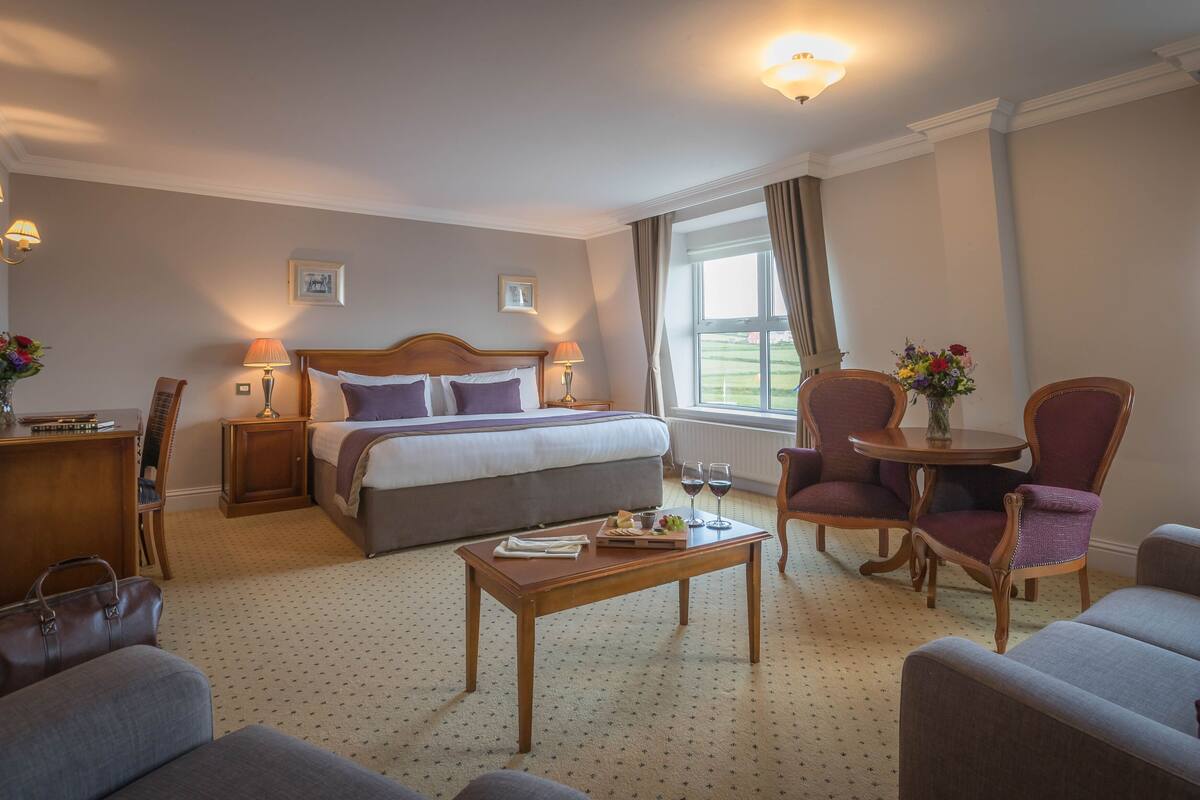 A hotel room with a bed, two chairs, a small table, a desk, and a window with curtains.