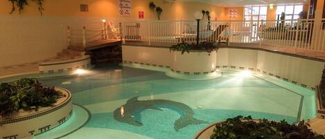 Une grande piscine intérieure ornée d’une fresque représentant des dauphins, entourée de rampes et d’escaliers blancs menant à un balcon.