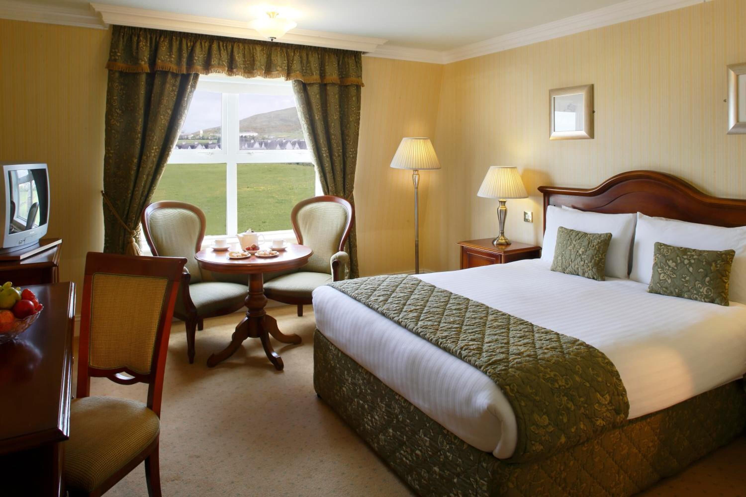 A hotel room with a large bed, two chairs, a small table, a TV, and a window with curtains.