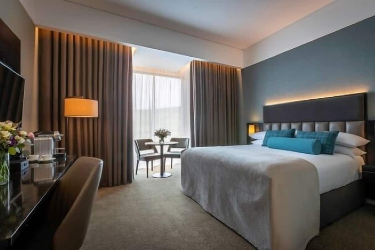 A hotel room with a large bed, a desk with a lamp, a chair, and a small table with a vase of flowers.