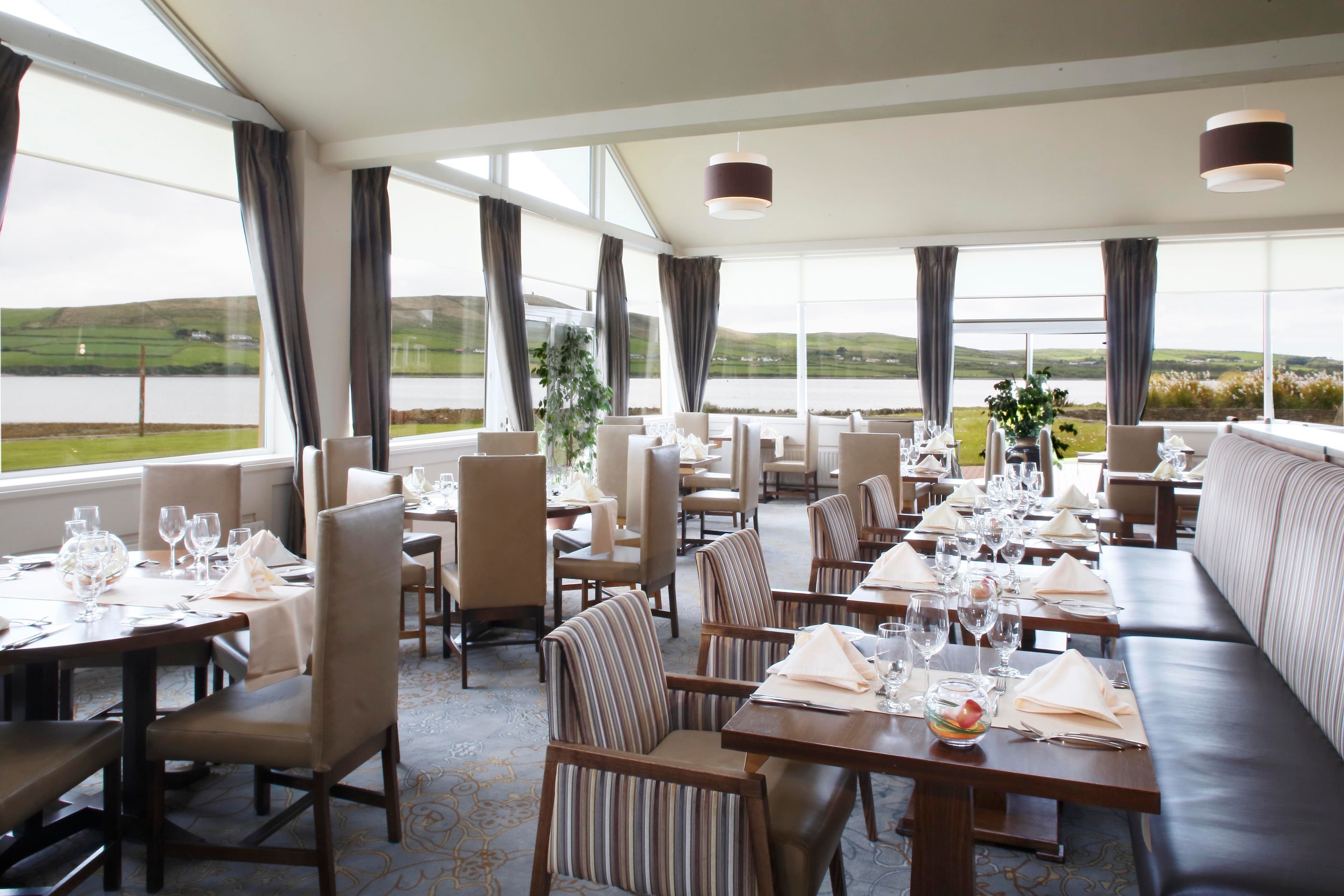 A restaurant with tables set for dining, large windows with curtains, and a view of the landscape.