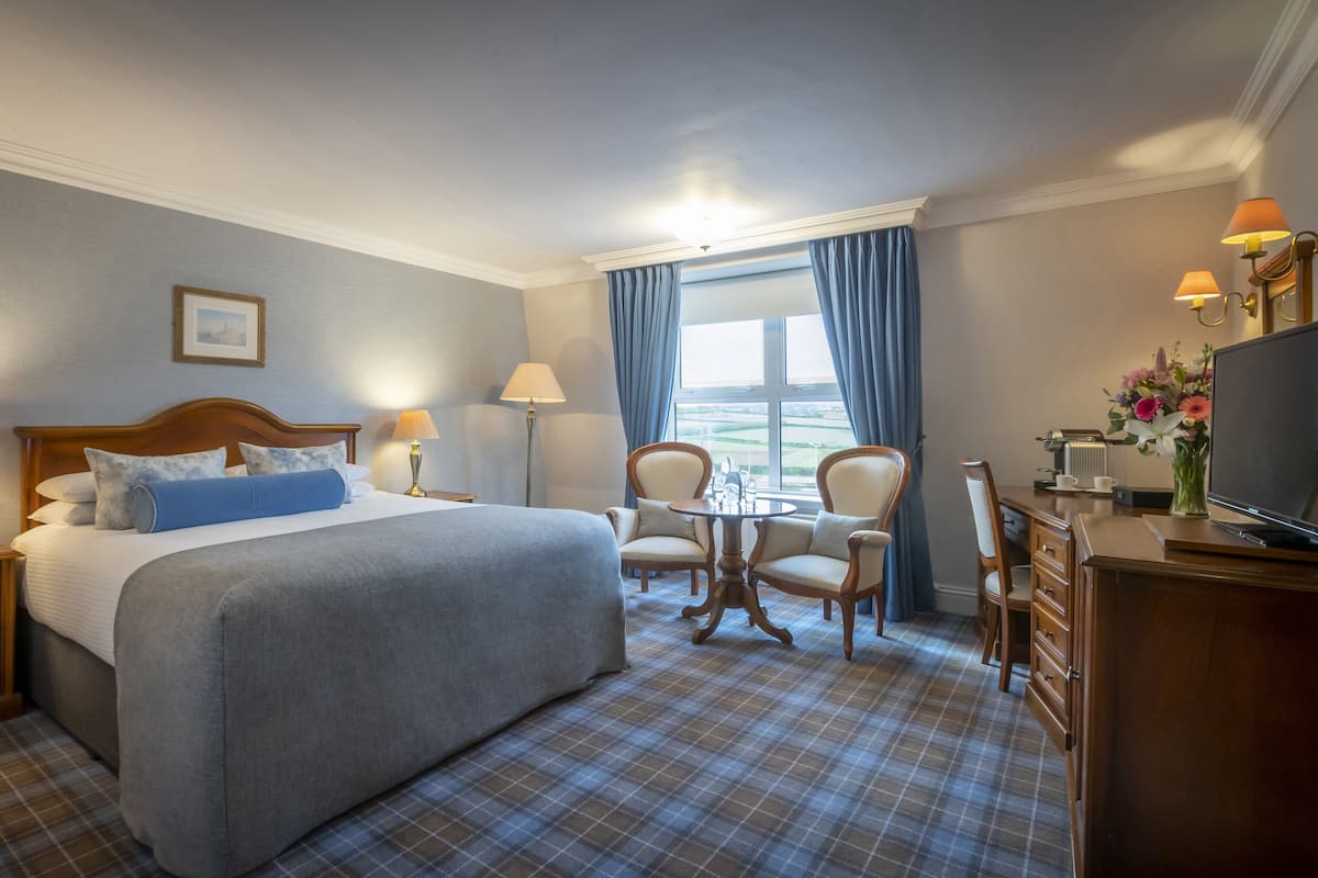A hotel room with a bed, two chairs, a desk with a TV, a coffee maker, and a window with curtains.